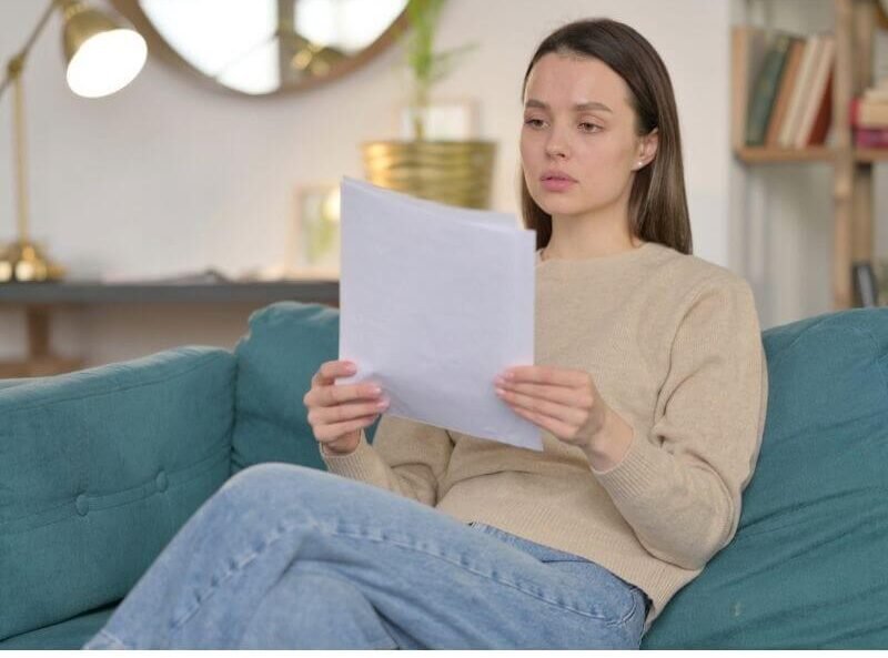 Mujer joven sentada en el sofá de su sala leyendo con atención un documento de Seguro Vida Mujer Banorte para revisar sus coberturas.