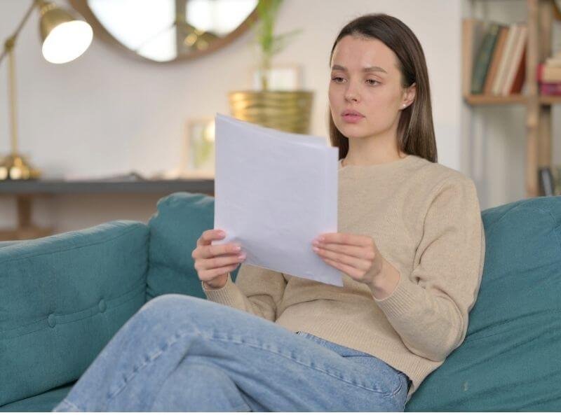 Mujer joven sentada en el sofá de su sala leyendo con atención un documento de Seguro Vida Mujer Banorte para revisar sus coberturas.
